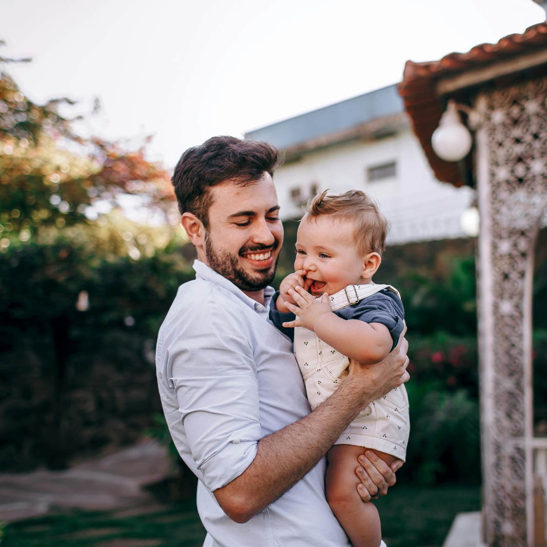 Man holding a child outdoors with a house and garden in the background