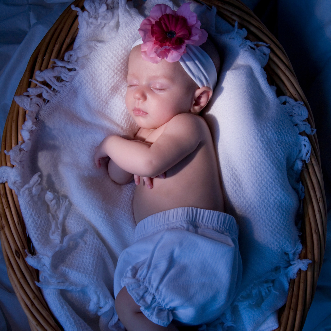 Newborn baby sleeping in a basket with white blankets and a pink flower headband.